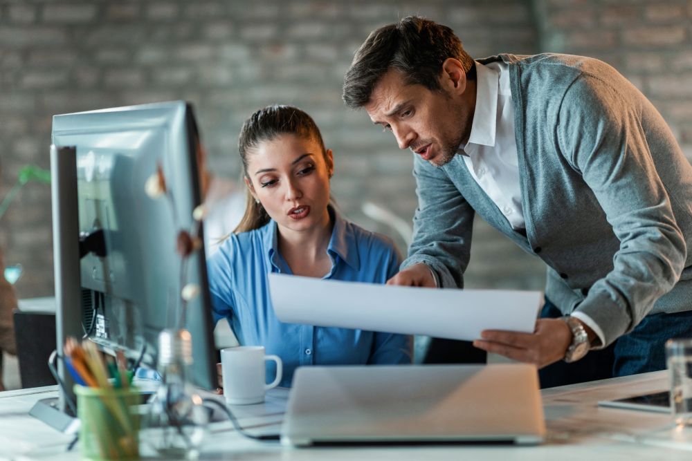 Dois profissionais analisam documento no computador enquanto discutem como explicar termos técnicos de TI.